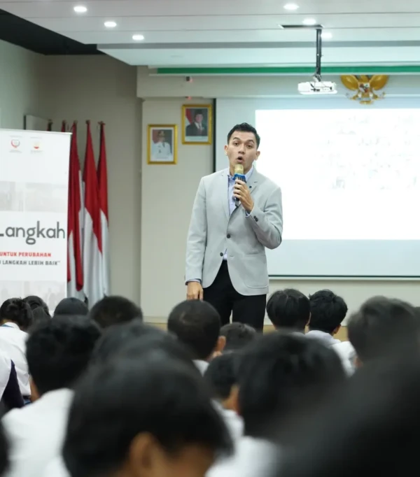 Seorang pembicara memberikan presentasi di depan audiens pelajar, dengan backdrop bendera dan banner program.
