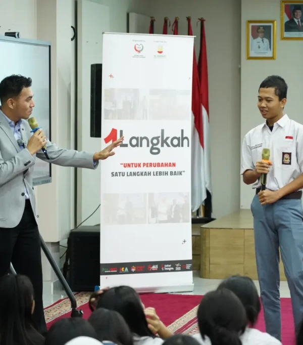 Acara motivasi di sekolah dengan pembicara dan siswa, banner "Satu Langkah Lebih Baik", semangat perubahan positif.