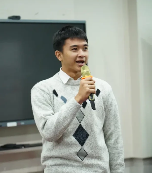 Pria berkemeja putih dan sweater abu-abu berbicara di depan audiens dengan mikrofon di tangan.