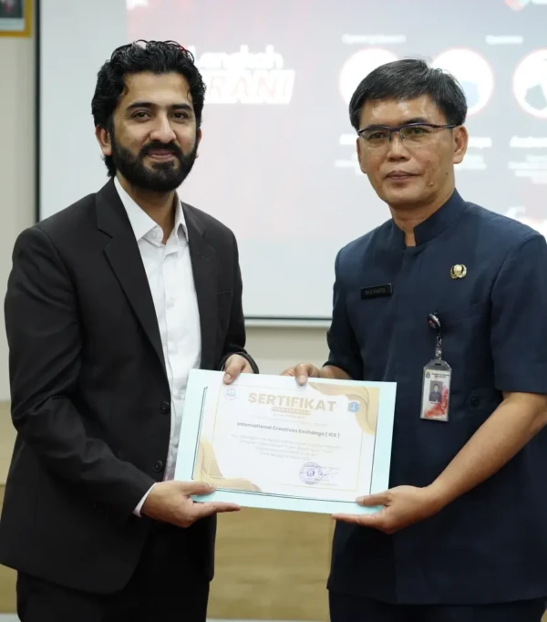 Dua pria menerima sertifikat dalam acara pertukaran internasional, dengan latar belakang presentasi di ruang konferensi.