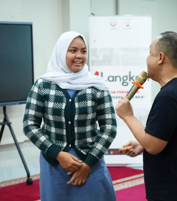 Seorang wanita berkerudung tersenyum saat berbicara dengan pembicara di acara presentasi. Latar belakang bendera Indonesia.